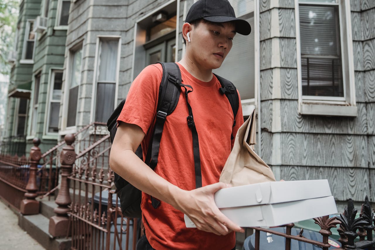 Courier delivering food in an urban neighborhood, holding pizza boxes and a brown paper bag.